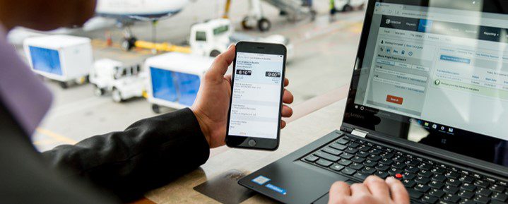 person sitting in airport terminal next to window overlooking the tarmac; their left white hand holds a cell phone and their right hand is on the mouse pad of a laptop with Concur displayed on the screen